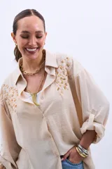 Camisa color beige con bordado de lentejuelas doradas en los hombros, corte clásico, cuello camisero y mangas largas con puños abotonados.