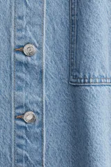 Camisa celeste de denim, corte amplio, con cuello clásico, manga corta, cierre frontal con botones metálicos, bolsillo de parche en el pecho y canesú en la espalda.