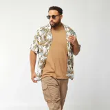Camisa de manga corta con estampado de hojas en tonos verdes, marrones y celestes sobre fondo blanco.