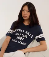Remera azul marino de algodón con cuello en V y mangas cortas con rayas blancas. Estampa frontal con texto "Beverly Hills Golf Club 1997 Local League" en blanco.