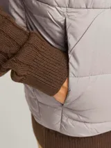 Chaleco puffer gris con cuello alto y cierre frontal con cremallera. Tiene bolsillos laterales y diseño acolchado horizontal.