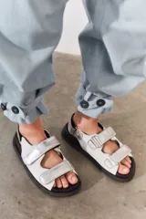 Sandalias de cuero color blanco con doble tira plateada con velcro, base negra y hebilla plateada.