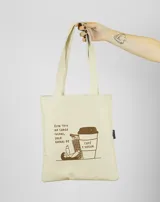 Bolso de mano tipo tote bag color crudo con asas largas y estampado con texto y dibujo de una persona recostada sobre una taza de café.