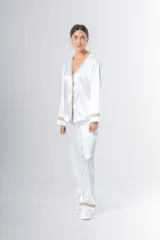 Pijama blanco de satén elastizado, compuesto por una camisa de manga larga con escote en V, abotonada adelante y con puntilla en escote y puños, y un pantalón recto con bolsillos laterales, elástico en la cintura y puntilla en el ruedo.