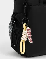 Cartera tipo tote de tela deportiva color negro, con correa regulable desmontable, manijas superiores, charm lateral con la palabra "Vibes", cierre superior, bolsillo frontal y bolsillos laterales de red.
