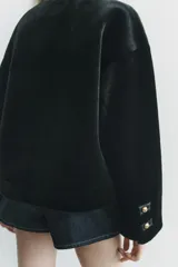 Chaqueta corta de cuello redondo y manga larga en tejido de pelo sintético color negro. Cierre frontal con botones metálicos dorados y bolsillos de plastrón. Colección Zara Woman.