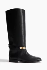 Botas altas de caña recta, color negro, con punta redondeada y taco bajo cuadrado. Presentan una correa decorativa alrededor del tobillo con una hebilla metálica rectangular de color dorado.