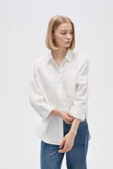 Camisa blanca de corte holgado, con cuello clásico, cierre frontal con botones, mangas largas con puños abotonados, dos bolsillos de parche en el pecho y bajo asimétrico.