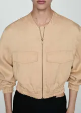 Chaqueta bomber color beige con cierre frontal, cuello redondo, mangas largas con puños elásticos y dos bolsillos delanteros con solapa.