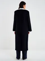 Abrigo largo negro de corte recto, con cierre frontal de un solo botón. Presenta cuello y puños blancos contrastantes, simulando una camisa debajo. Tiene bolsillos delanteros con solapa y abertura posterior.