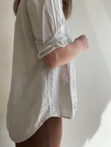 Blusa color crema con cuello camisero, mangas cortas con dobladillo y cierre frontal con un botón de madera. Presenta bordados con motivos de paisley y perforaciones.