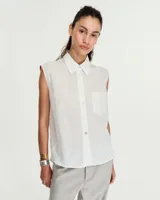Camisa blanca sin mangas en voile de algodón liviano y fresco, con fit relajado, cuello camisero, cartera con botones de nacar y un bolsillo plaqué en delantero. Las sisas están envivadas internamente con una tela contratono. Tiene costura en centro espalda y recorte en cintura trasera con tajo que se superpone. El ruedo es escalonado, más corto adelante. Largo a la cadera.
