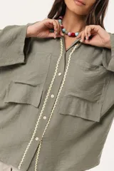 Camisa verde militar de manga larga con silueta oversize, dos bolsillos delanteros y corte levemente más largo en la espalda. Escote en V abotonado con detalle de galón picot a lo largo de la cartera y botones a tono. Isotipo bordado en la parte trasera.