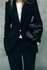 Blazer negro largo con cuello solapa, manga larga con hombreras, bolsillos delanteros de vivo y cierre frontal con un botón.
