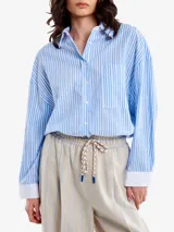 Camisa celeste y blanca a rayas verticales, de corte holgado, con cuello clásico, bolsillo en el pecho, mangas largas con puños blancos y cierre frontal con botones.