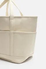 Bolso tipo shopper color crema, fabricado en tejido de algodón. Tiene doble asa de mano, compartimento principal con cierre de cremallera, seis bolsillos planos interiores, funda interior para dispositivos electrónicos de hasta 13 pulgadas con cierre de cremallera, bolsillo plano frontal y bolsillos planos laterales.