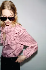 Camisa de manga larga color rosa claro con finas rayas verticales en tono rosado más oscuro. Presenta cuello mao y un gran volante fruncido que recorre el frente, desde los hombros hasta el centro. Los puños son elásticos.