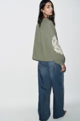 Blusa verde oliva de algodón con cuello redondo y escote en pico con cordones y borlas. Tiene manga larga con bordados florales en los hombros, jaretas y entredoses delanteros. Cierre frontal con botones.