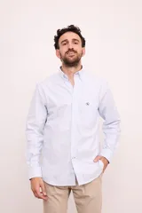 Camisa celeste y blanca a rayas verticales, de corte clásico, manga larga y cuello abotonado. Tiene bolsillo en el pecho con logo bordado.
