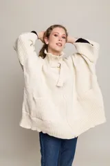 Tapado tejido de punto grueso color crudo, con cuello alto, cierre frontal con cremallera y hebilla decorativa, mangas amplias y bolsillos delanteros.