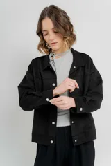 Chaqueta color beige de corte recto, estilo camisa, confeccionada en gamuza con elastano. Presenta cuello camisero, cierre frontal con botones metálicos, bolsillos grandes en el frente y charreteras en los hombros.