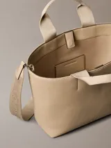 Mini cartera tote Calvin Klein color beige, confeccionada en poliuretano, con doble asa de mano y correa de hombro ajustable extraíble. Incluye cierre con botón a presión y bolsillo interior para tarjeta.