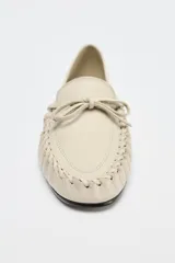 Zapato plano tipo mocasín color crema, confeccionado en cuero, con detalle de lazo con pasador y fruncido en la parte delantera. Pespunte en el corte y acabado en punta redonda.