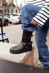 Bota negra de cuero con caña media, suela track, correa con hebilla en el tobillo y arnés con argolla metálica.