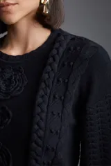Sweater negro de punto con cuello redondo y mangas largas. Presenta tres flores tejidas en el frente y detalles de trenzas verticales.