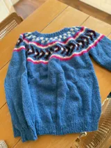 Sweater tejido a mano en lana mohair color crudo, con cuello redondo y mangas largas. Presenta un diseño con motivos geométricos en tonos marrón y verde en la parte superior del pecho.