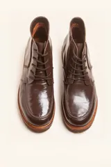 Bota de cuero vacuno con acabado charol, color marrón oscuro. Presenta un diseño de caña corta con cordones frontales y costuras decorativas en la puntera. La suela es de goma tipo crepe.