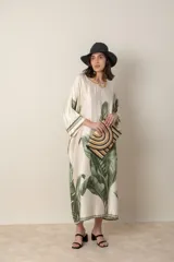 Vestido largo color natural con estampado de hojas de palmera en tonos verdes. Tiene corte amplio tipo túnica, cuello redondo, mangas tres cuartos, bolsillos laterales y ruedo con ribete.