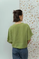 Blusa de lino color azul claro con escote en V cruzado y manga corta. Se ajusta en el centro del busto con un botón redondo de madera o metal con relieve.