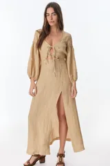 Vestido color beige de lino, con escote en V, mangas largas abullonadas y lazos frontales. Tiene corte a la cintura y falda larga con tajo frontal.