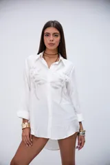 Camisa oversize color crudo con cuello clásico, cierre de botones frontales y mangas largas con puños. Presenta bordados florales con lentejuelas y mostacillas plateadas en la parte delantera.