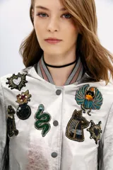 Campera bomber de cuero ovino color plateado, con cuello, puños y ruedo de ribb metalizado. Presenta apliques de parches bordados con lentejuelas y mostacillas.