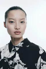 Camisa de sarga estampada con flores en blanco y negro, de corte recto, con cuello clásico, cierre frontal con botones, mangas largas con puños abotonados y bajo redondeado.