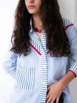 Camisa celeste a rayas verticales blancas y azules, con bolsillo de parche en el frente y terminaciones en crochet rojo en cuello, mangas y hombros.