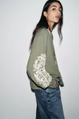 Blusa verde oliva de algodón con cuello redondo y escote en pico con cordones y borlas. Tiene manga larga con bordados florales en los hombros, jaretas y entredoses delanteros. Cierre frontal con botones.