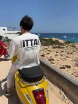 Remera color crema de corte recto y mangas cortas, con estampado en la espalda con el logo "ITTAI" en negro con líneas rojas, azules y blancas, y la leyenda "AN IBIZA PRODUCE. Ittai Stars Department Gallery - Ibiza Edition. If you're in Ibiza, 112 is not an option.".