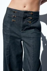 Pantalón culotte de tiro alto en denim azul oscuro, con pernera ancha y corta terminada en vuelta. Presenta falsos botones dorados en la parte delantera, bolsillos frontales y cierre con cremallera y gancho.