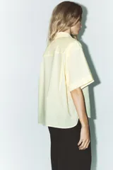 Camisa color crema de cuello solapa y manga corta con vuelta. Tiene un bolsillo de parche en el delantero, bajo asimétrico con aberturas laterales y cierre frontal con botones.