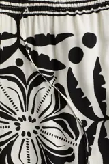 Pantalón largo de viscosa color blanco con estampado de flores y figuras geométricas en negro. Tiene tiro alto, cintura elástica con cordón ajustable, bolsillos laterales y corte holgado.