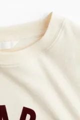 Remera color crema de corte holgado, con cuello redondo y estampado frontal con la frase "Le Marais Paris" en color borgoña.