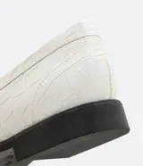 Zapato mocasín de color blanco con textura de piel de cocodrilo y detalle de borla frontal. Posee punta redonda, tacón plano y suela en contraste de color negro.