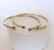 Pulsera rígida abierta en color dorado, con iniciales grabadas en los extremos.