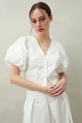 Blusa blanca con lunares grises, escote en V, mangas cortas abullonadas y cierre frontal con botones. 