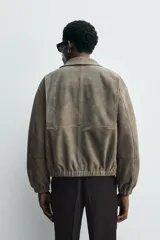 Cazadora de hombre color verde oliva, confeccionada en piel con acabado ante. Presenta un corte boxy fit, cuello solapa, cierre frontal de cremallera, bolsillos delanteros y elásticos en puños y bajo.