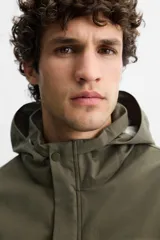 Parka verde militar confeccionada en tejido técnico ligero resistente al viento, con cuello alto, capucha, manga larga con puños ajustables, bolsillos de plastrón con solapa en la cadera, bolsillo interior, bajo ajustable con elásticos y cierre frontal con cremallera oculta por solapa con botones a presión.