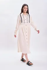 Vestido largo de muselina color crudo con bordado negro en el cuello y la pechera, corte evasé, manga larga y abotonadura frontal.